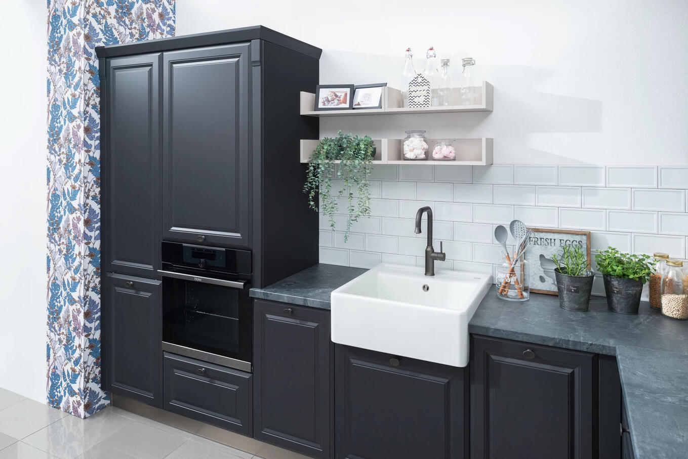 Elegantná kuchyňa s čiernymi skrinkami a kontrastným bielym obkladom, ktorá prináša sofistikovaný a moderný štýl. Tmavá kuchyňa s čiernymi skrinkami, veľkým keramickým drezom a bielym obkladom, doplnená otvorenými poličkami s dekoráciami.